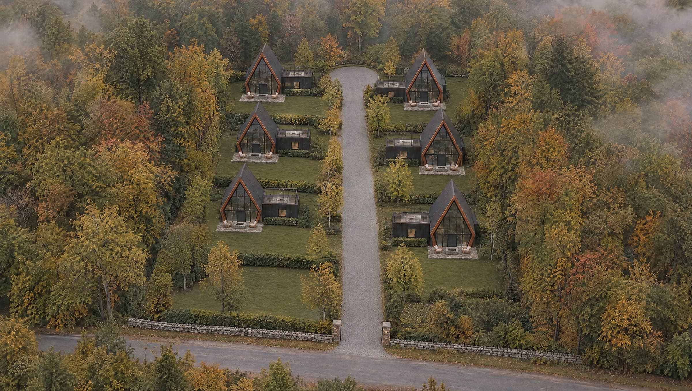 The Avara Residences from the air in early autumn: a straight gravel lane runs in from a fieldstone gateway to a turning circle, with three black A-frame houses on each side, each with a fieldstone patio, a stone fire table and lounge chairs in front of its glass gable, lawns and hedges between them, all within mature woodland