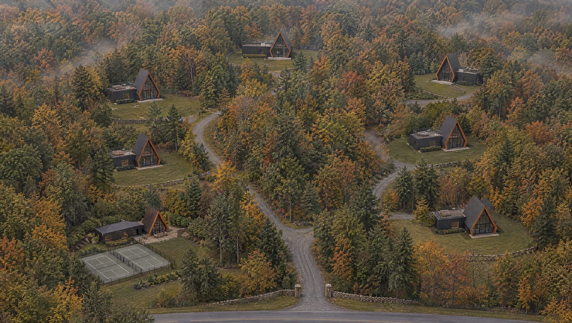 The Avara Residences from the air in early autumn: a private lane enters through a fieldstone gateway and divides around the woods to six black A-frame houses, each in its own clearing with a lawn and old stone walls, and a small A-frame clubhouse with two pickleball courts near the entrance