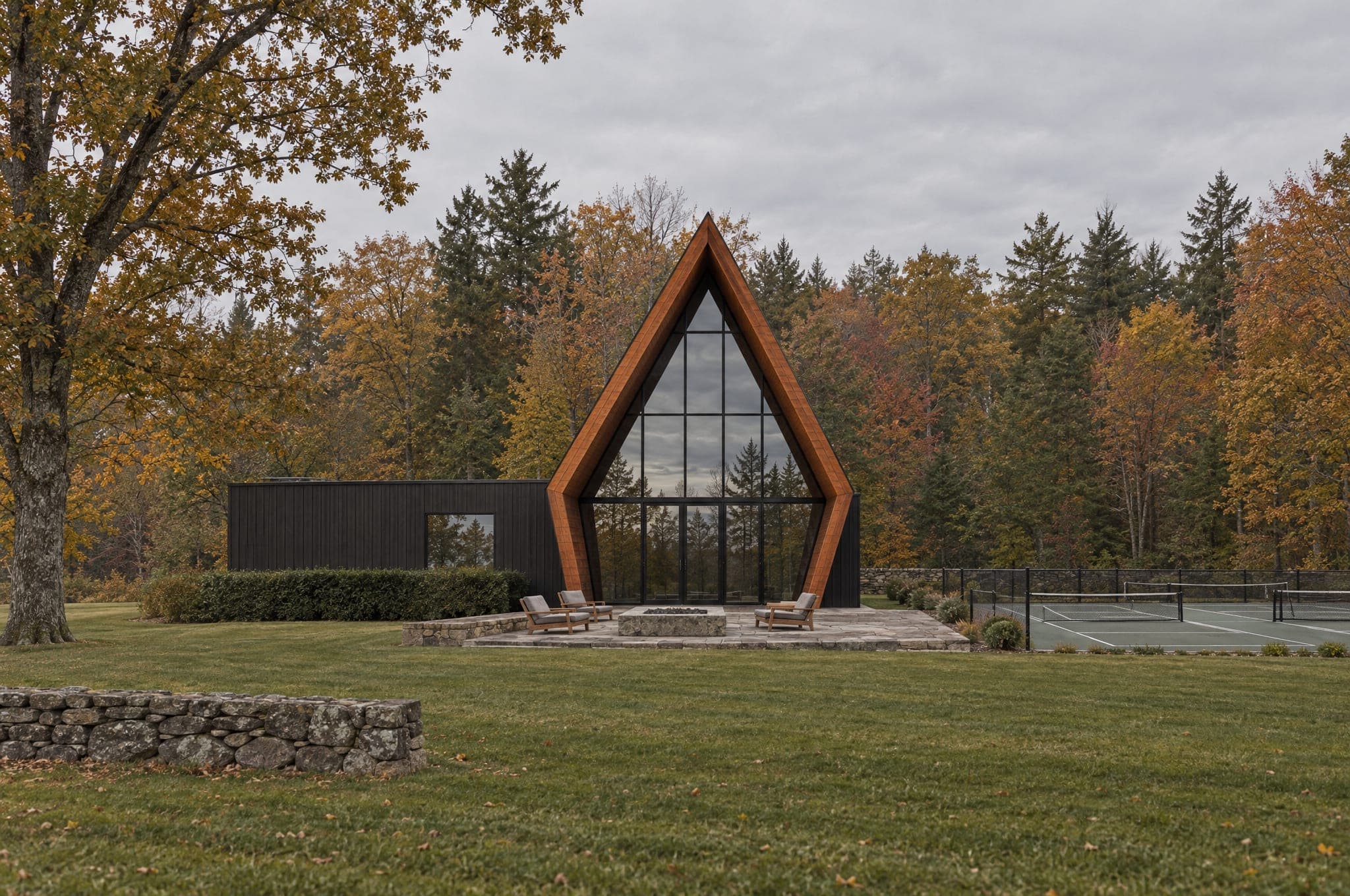 The residents' clubhouse: a small black A-frame with a glass gable under a wood-trimmed porch shell and a low black wing, a fieldstone terrace with lounge chairs and a fire table, and the pickleball courts beside it at the edge of autumn woods